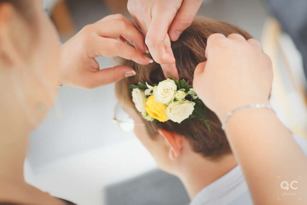 Gros plan sur les mains de la coiffeuse arrangeant une couronne de roses blanches et jaunes dans les cheveux d'une mariée. Les cheveux bruns de la mariée sont rassemblés en un chignon élégant.