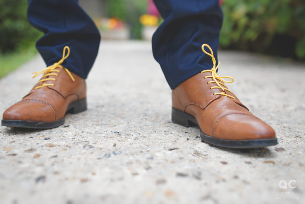 Une vue rapprochée de chaussures en cuir marron à lacets jaunes sur un chemin en béton. Les chaussures sont portées par le marié vêtu d'un pantalon bleu marine.