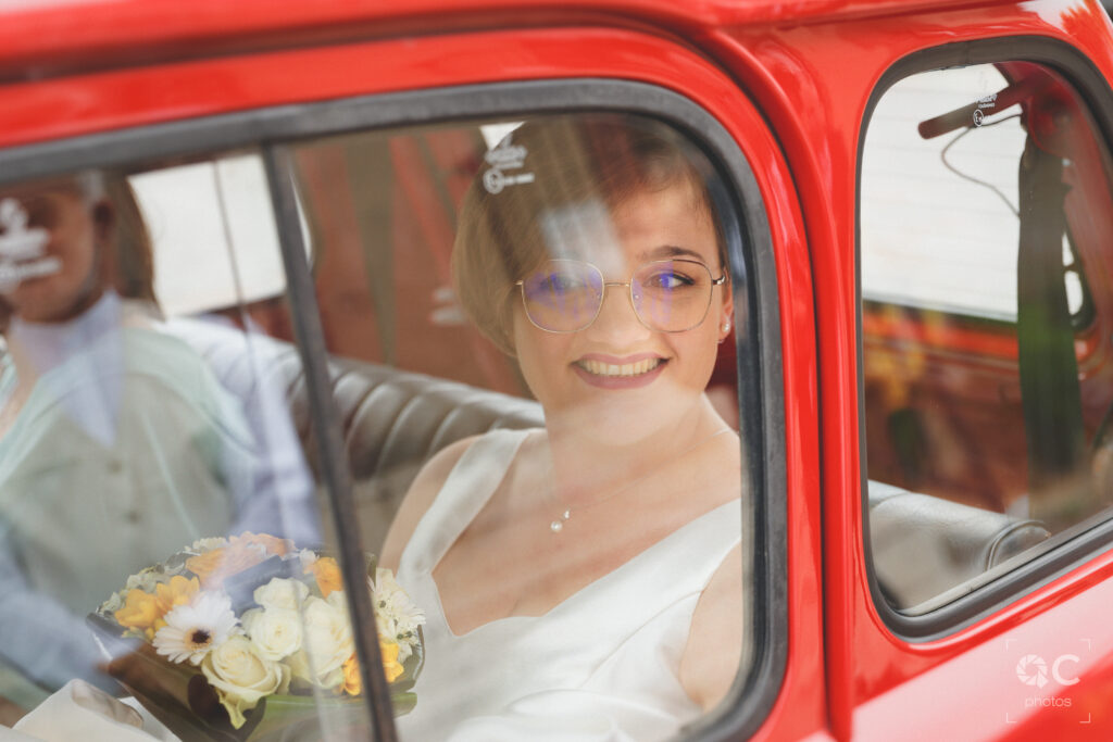 Vue rapprochée d'une mariée souriante assise dans une voiture rouge classique. Elle porte une robe blanche et tient un bouquet de fleurs. Elle regarde vers la droite hors de la voiture.