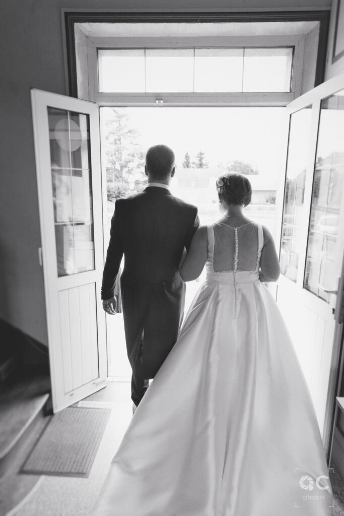 Vue arrière d'un couple marié en noir et blanc quittant la mairie. Ils s'éloignent, le regard tourné vers l'avenir.