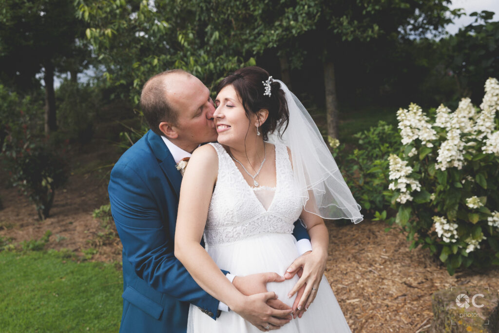 Un couple marié, s'embrasse tendrement dans un jardin. La mariée est enceinte et les mains du couple forment un cœur sur son ventre. Des fleurs blanches sont visibles en arrière-plan.
