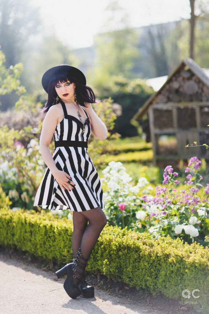 Photo d’une jeune femme portant une robe rayée noir et blanc et un chapeau noir, posant dans un jardin fleuri.