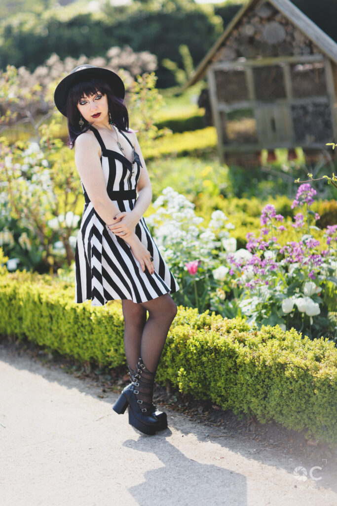 Une femme élégante portant une robe rayée noire et blanche, un chapeau noir et des bottines à plateforme, pose dans un jardin fleuri. Un petit chalet en bois se trouve en arrière-plan.