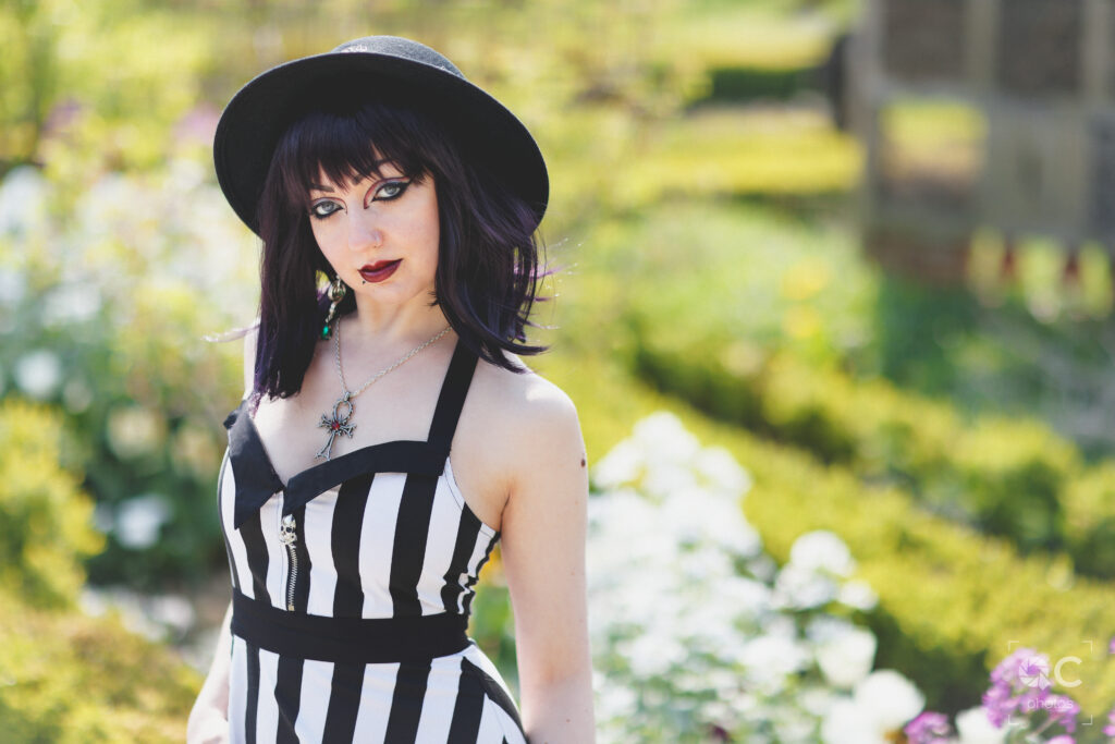 Une jeune femme aux cheveux noirs et aux yeux bleus vifs, portant une robe rayée noir et blanc et un chapeau noir, pose dans un jardin. Des fleurs et des buissons verdoyants l’entourent, créant un contraste saisissant entre l'élégance gothique de la modèle et la beauté de la nature. La scène est baignée de lumière naturelle.