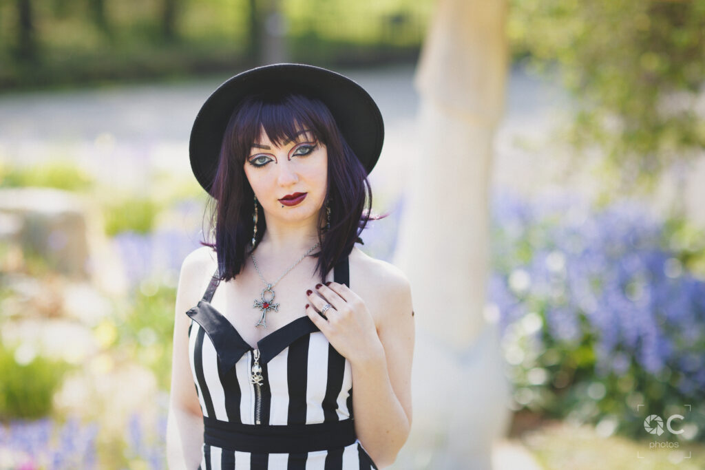 Portrait d'une jeune femme aux cheveux violets portant un chapeau à larges bords et une robe rayée noir et blanc, dans un jardin fleuri. Elle porte un collier avec un ankh et une bague. Style gothique romantique.