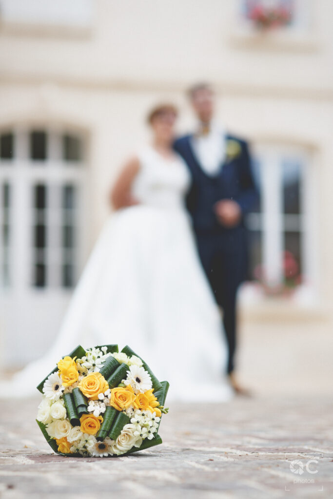 Bouquet de mariée sur un sol en pierre. Les mariés flous sont à l'arrière-plan. Les fleurs sont jaunes et blanches.