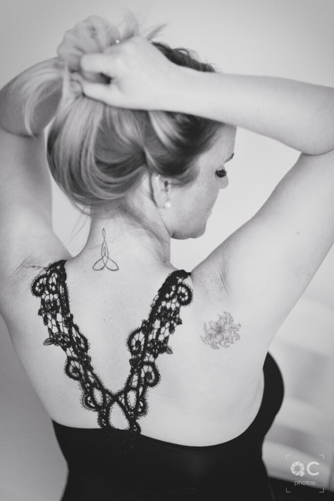 Photo en noir et blanc d'une femme vue de dos, ses cheveux attachés en chignon. Elle porte un soutien-gorge en dentelle noire, révélant de délicats tatouages sur son dos et ses épaules.