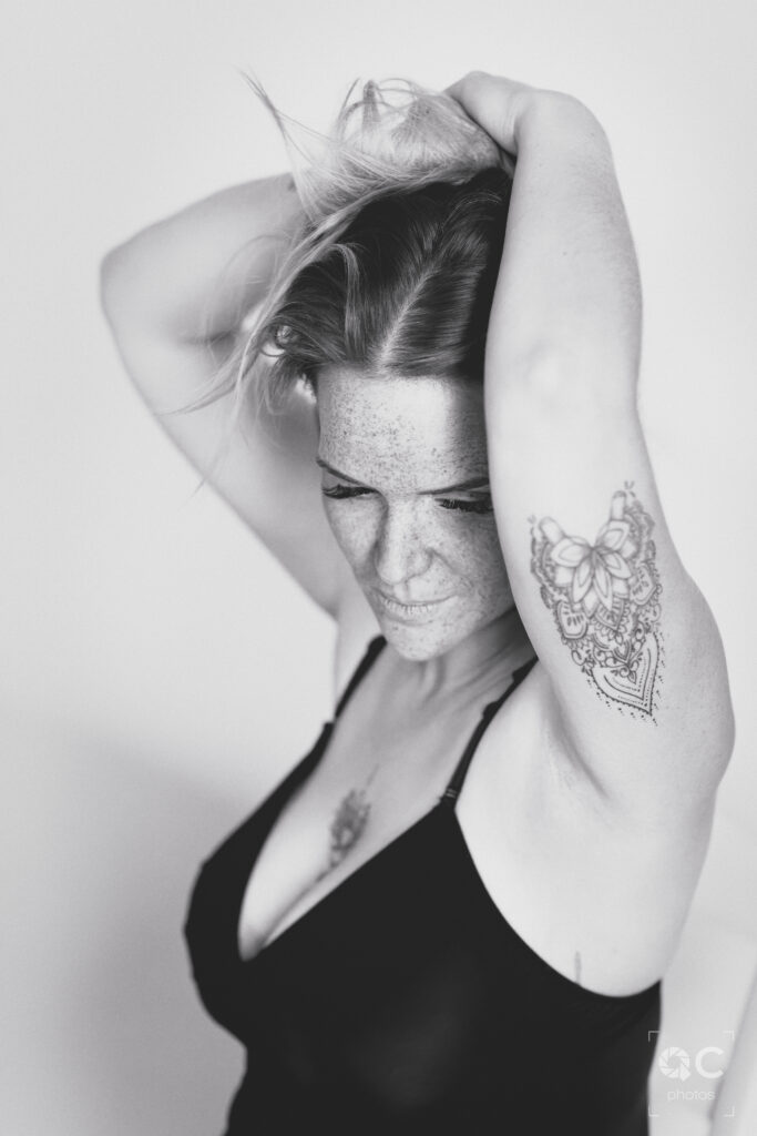 Femme aux cheveux mi-longs et aux taches de rousseur, portant un soutien-gorge noir. Ses bras sont levés, elle se tient la tête. Un tatouage ornemental orne son bras. L’ambiance est intime et puissante.