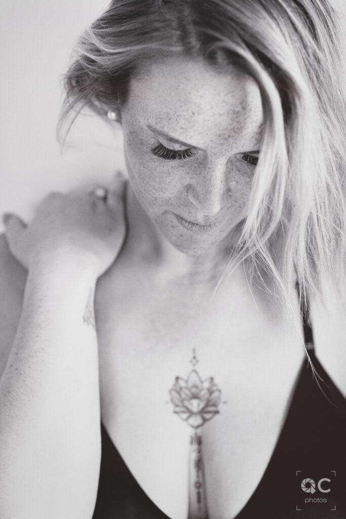 Photo en noir et blanc d'une femme aux cheveux blonds, légèrement penchée en avant. Elle porte un soutien-gorge noir et dévoile un tatouage délicat en forme de lotus sur sa poitrine. L'image est à la fois intime et artistique, mettant en lumière la beauté naturelle du modèle.