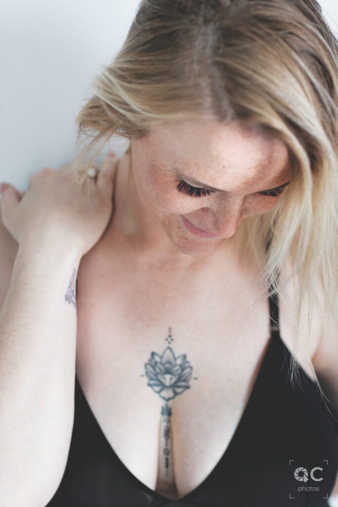 Une femme blonde aux taches de rousseur, portant un soutien-gorge noir, se tient les épaules. Elle porte un tatouage de lotus sur la poitrine. L'image est à la fois intime et élégante.