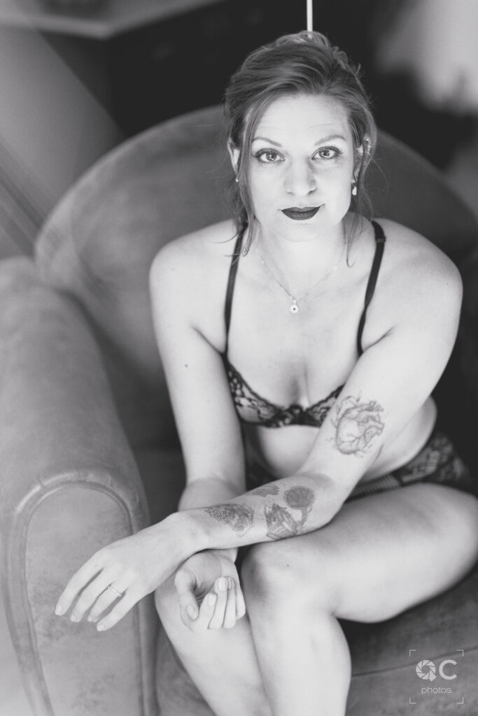 Photo en noir et blanc d’une femme assise sur un fauteuil, portant une lingerie délicate. Elle a plusieurs tatouages sur les bras et regarde directement l’objectif avec confiance. L’ambiance générale est sensuelle et élégante.