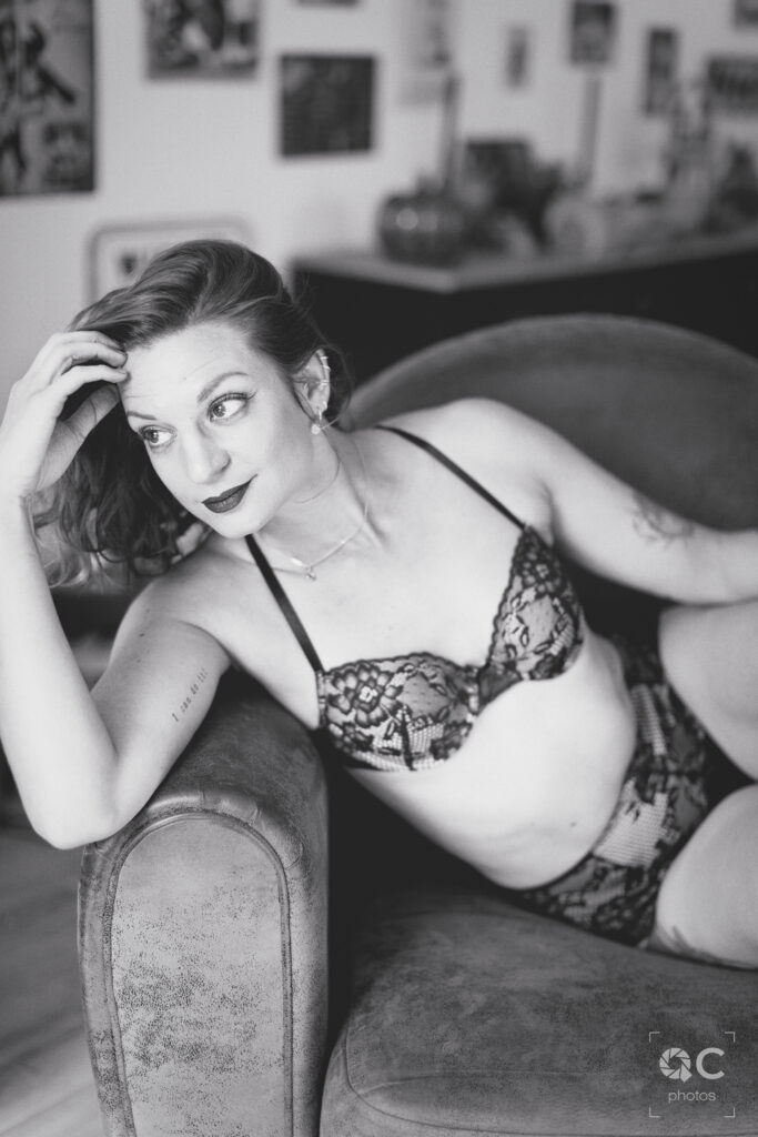 Photo en noir et blanc d'une femme aux cheveux roux, légèrement ondulés, assise sur un fauteuil. Elle porte une lingerie noire en dentelle et regarde au loin, une expression douce et pensive sur le visage. Son bras gauche repose sur le dossier du fauteuil, révélant un tatouage discret sur son avant-bras.