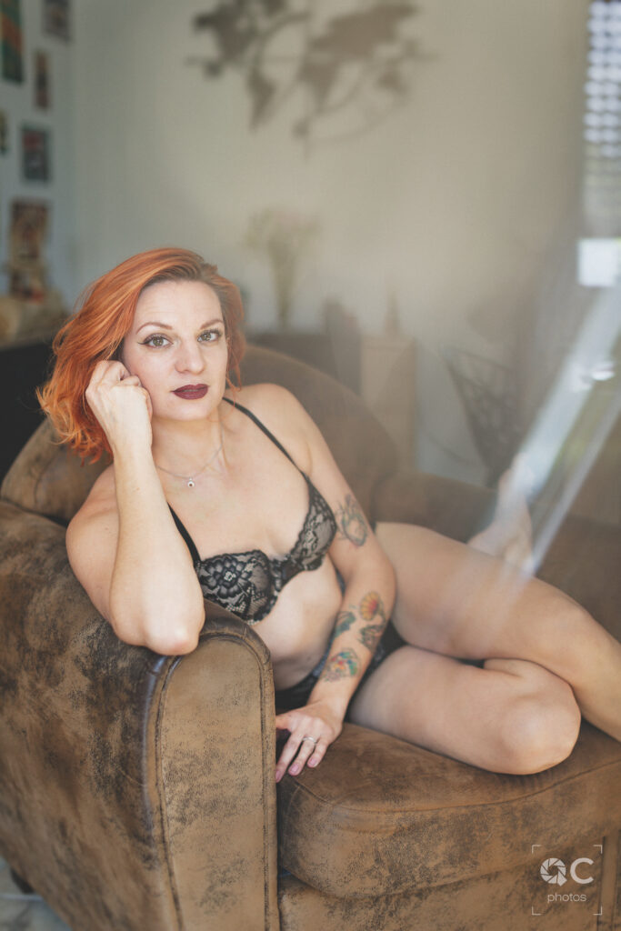 Portrait d'une femme rousse aux cheveux courts et aux tatouages sur les bras, assise sur un fauteuil en cuir vieilli. Elle porte un soutien-gorge en dentelle noire. L'image est douce et sensuelle, dans une ambiance chaleureuse et intime.