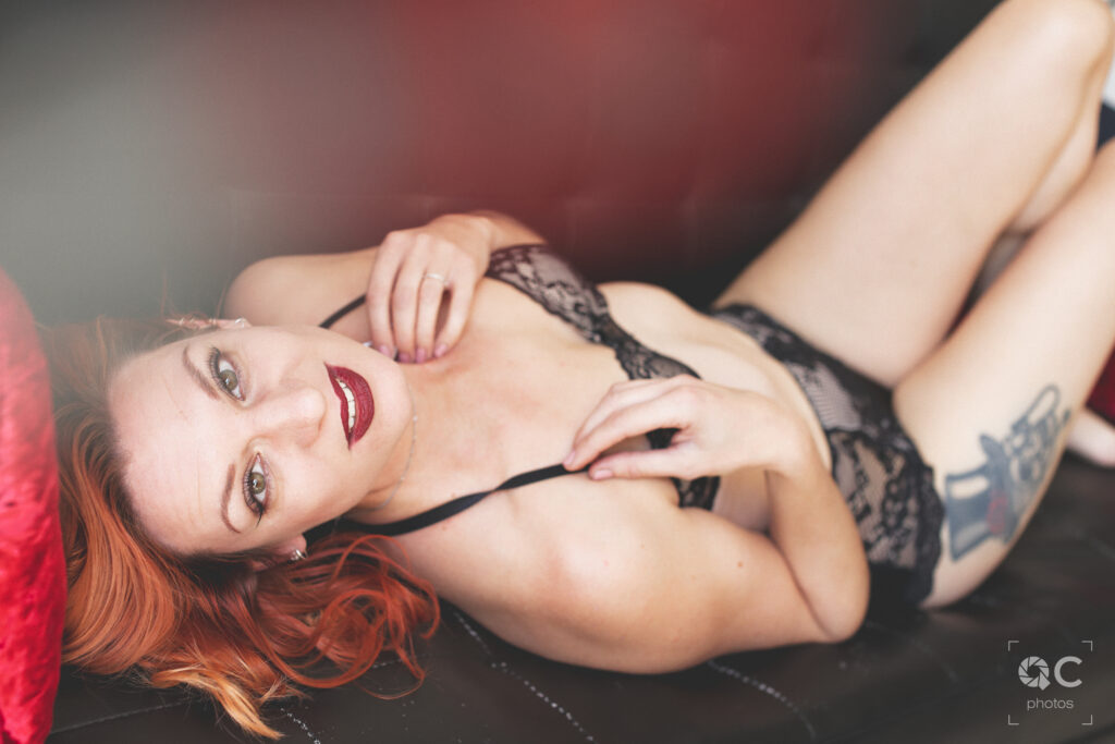 Une femme aux cheveux roux allongée sur un canapé, vêtue d’une lingerie noire en dentelle. Elle regarde la caméra avec un air de confiance et de sensualité. L’image est douce et mystérieuse, mettant en valeur la beauté naturelle de la femme.