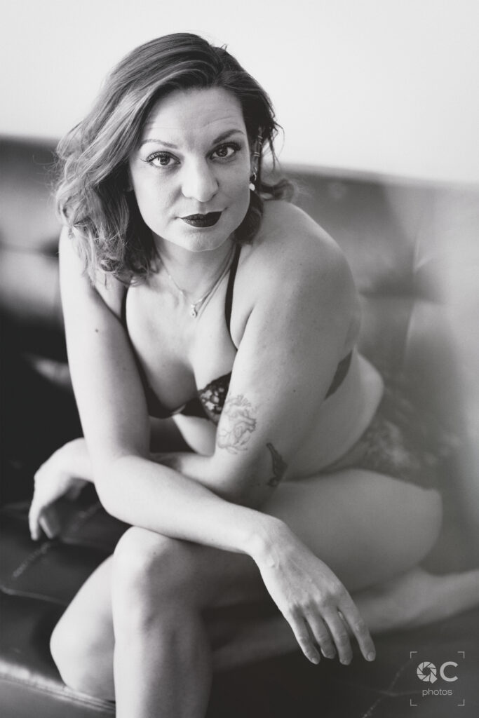 Portrait en noir et blanc d'une femme assise sur un canapé, vêtue d'une lingerie délicate. Elle regarde directement l'objectif avec assurance et sérénité. Ses bras sont croisés sur ses genoux, et sa posture est à la fois détendue et élégante.