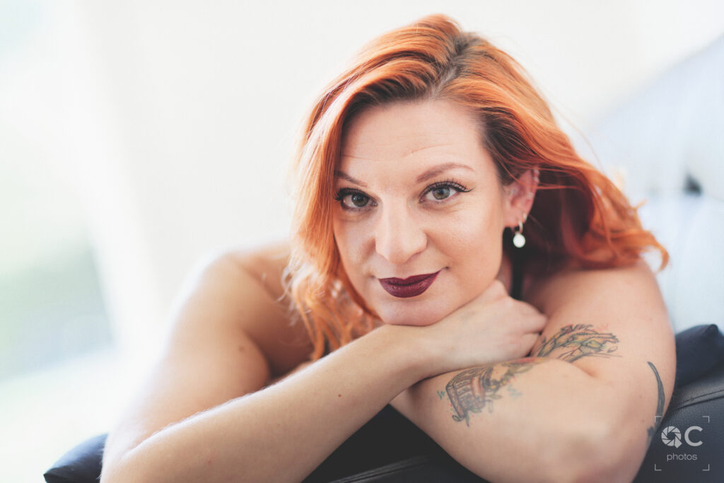 Portrait intime d'une femme aux cheveux roux, le visage légèrement penché, les bras croisés sur le torse, dévoilant un tatouage délicat sur son bras. L'ambiance est douce et sensuelle.